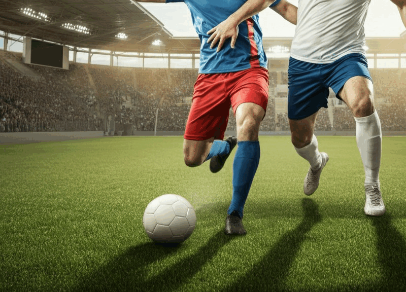 Due giocatori di calcio in azione sul campo, con gli spalti affollati di uno stadio sullo sfondo e un cielo pomeridiano chiaro, perfetto scenario per le scommesse Champions League.
