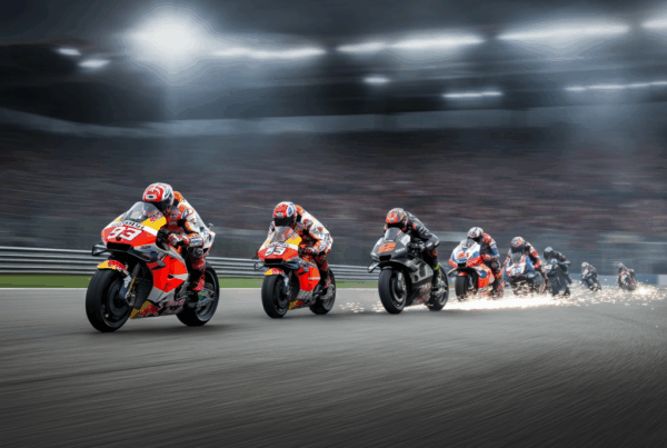 Motociclette da competizione che affrontano curve strette su un circuito professionale, simbolo della Classifica MotoGP dei Crash, con scintille che si sprigionano dalle pedane inclinate e una folla festante sullo sfondo.