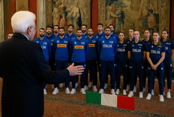 Il Presidente Sergio Mattarella, di spalle, riceve le Nazionali italiane di volley maschile e femminile in una sala elegante che ricorda il Quirinale. Gli atleti, in divisa azzurra con la scritta “Italia”, ascoltano attentamente mentre sul pavimento è visibile una piccola bandiera tricolore.
