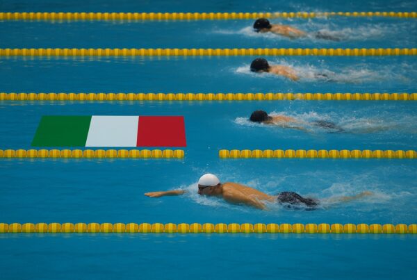 Vista dall’alto di una gara di nuoto in una piscina olimpionica, con un nuotatore italiano in netto vantaggio rispetto agli avversari. Nella sua corsia, davanti a lui, è visibile la bandiera dell’Italia nitida e piatta sull’acqua. Gli altri atleti seguono distanziati sullo sfondo. L’immagine celebra i nuotatori italiani famosi e la loro supremazia nelle competizioni internazionali.