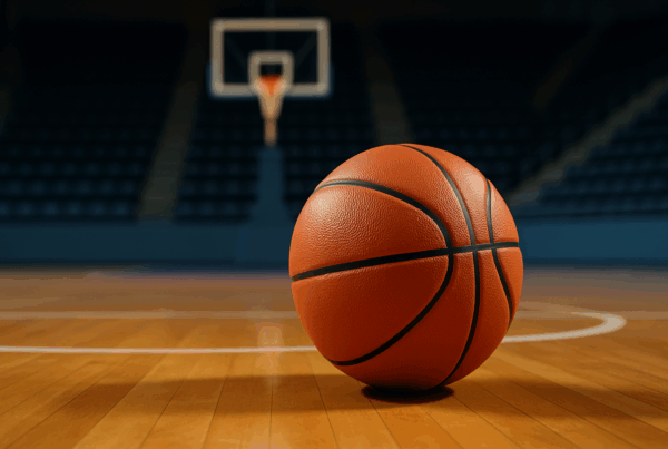 Un pallone da basket realistico posato sul parquet di un campo indoor con il canestro sfocato sullo sfondo.