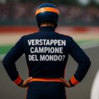 Pilota generico di Formula 1 visto di spalle su un circuito, con tuta blu e arancione e la scritta VERSTAPPEN CAMPIONE DEL MONDO? stampata in grande sulla schiena, mentre osserva la pista davanti a sé.
