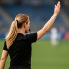 Arbitro donna di spalle su un campo da calcio, capelli biondi raccolti in una coda, braccio alzato e fischietto in bocca durante una decisione di gioco.
