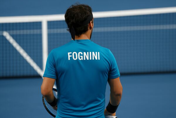 Fabio Fognini di spalle su un campo da tennis blu, in maglia azzurra con scritta “FOGNINI”, racchetta in mano e rete sullo sfondo. la foto fa riferimento all'intervista di Fognini a Belve