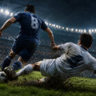 Tackle nel calcio durante una partita in notturna, con un difensore in scivolata che recupera il pallone e un avversario in corsa su un campo illuminato.