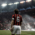 Franco Baresi di spalle con la storica maglia numero 6 del Milan osserva lo stadio illuminato, in un'illustrazione iperrealistica che rende omaggio alla leggenda del calcio italiano.