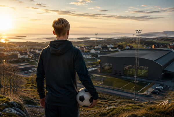 Erling Haaland Bryne, giovane calciatore di spalle con pallone davanti ai campi da calcio della città norvegese al tramonto.